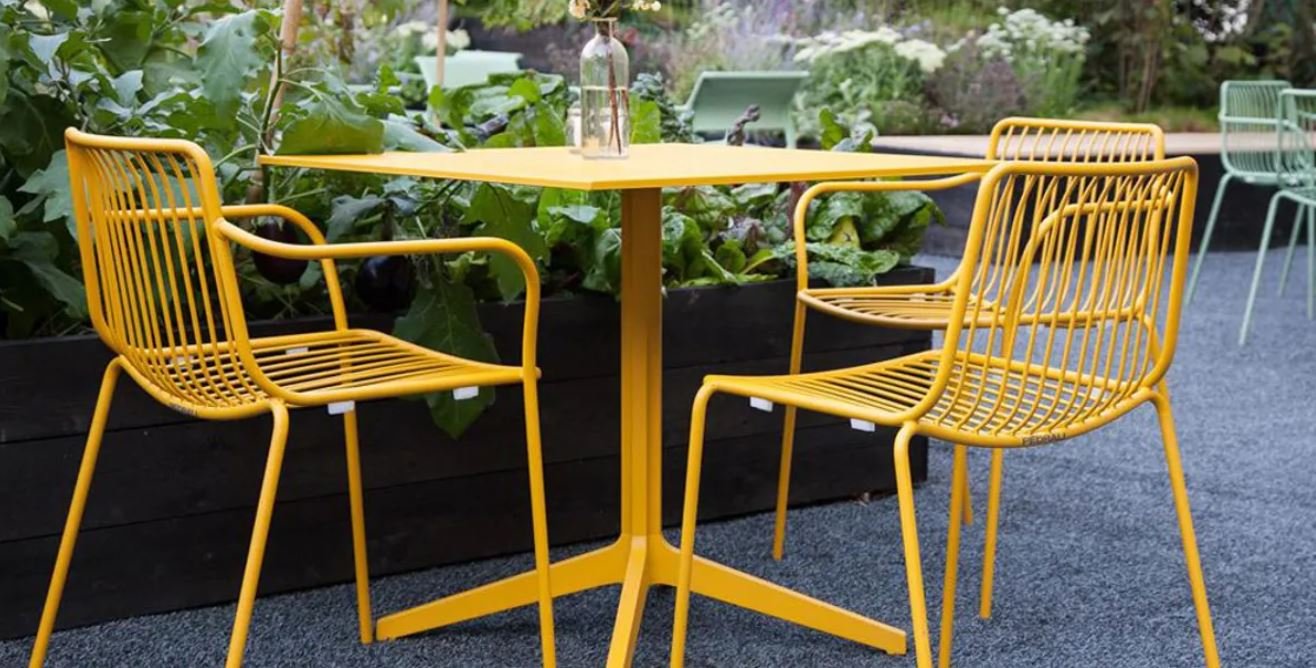 Four yellow Pedrali Nolita chairs with metal frames and slatted seats, arranged around a yellow square table in a garden with raised beds. 