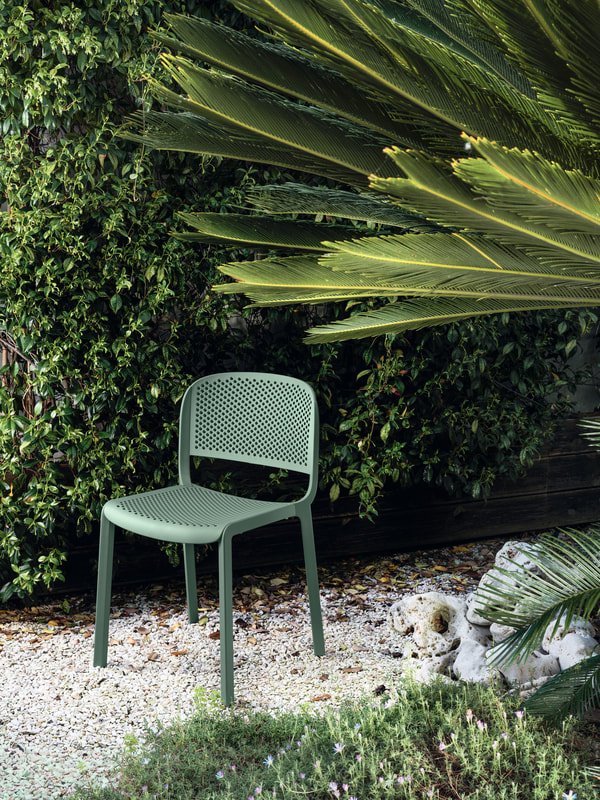Olive green Pedrali dome chair in polypropylene with perforated seat and back, placed in a garden with gravel and thick vegetation in the background